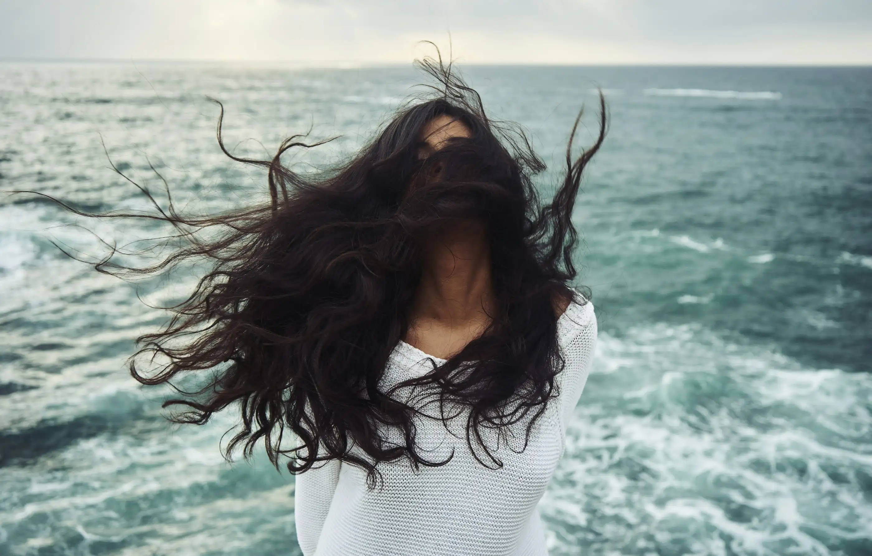 A woman's long dark hair blowing across her face in the wind beside a rough sea
