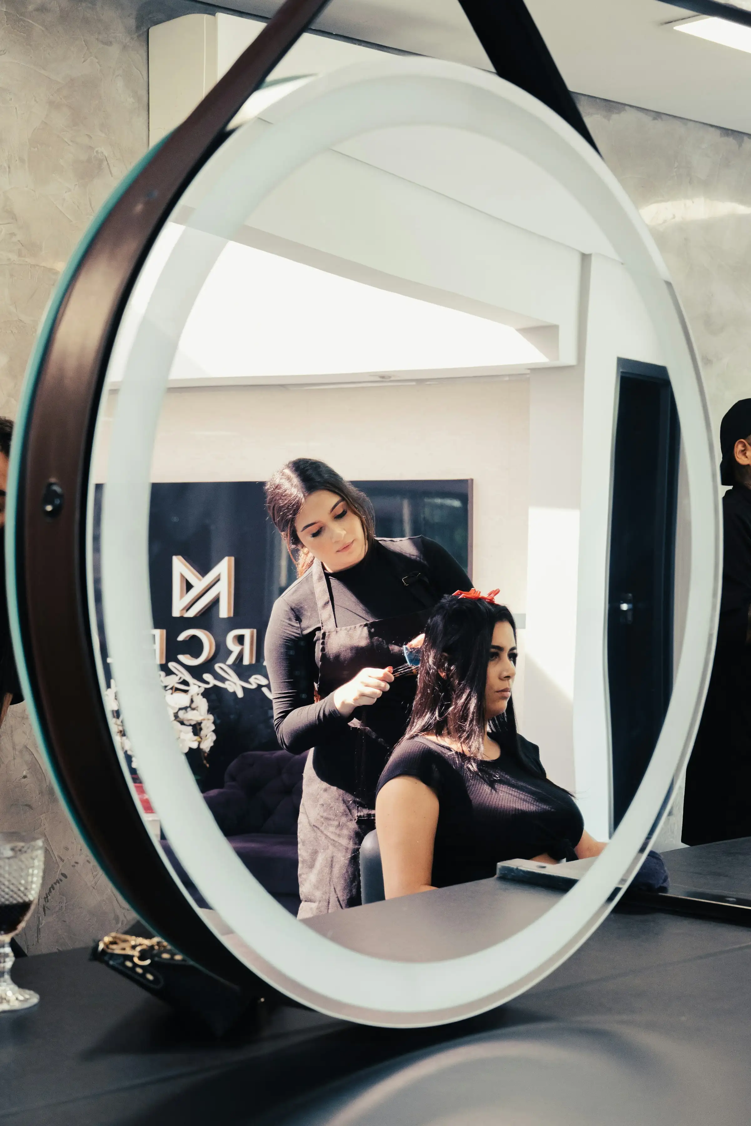 A stylist working on a client's hair, seen in a round salon mirror